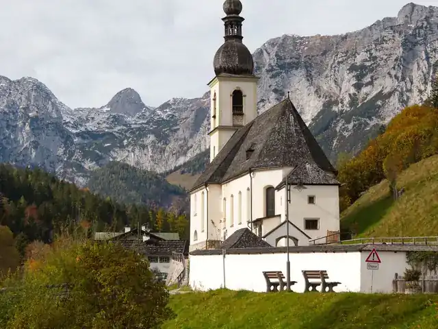 White Bavarian village church with an onion-dome tower below rocky mountains in autumn