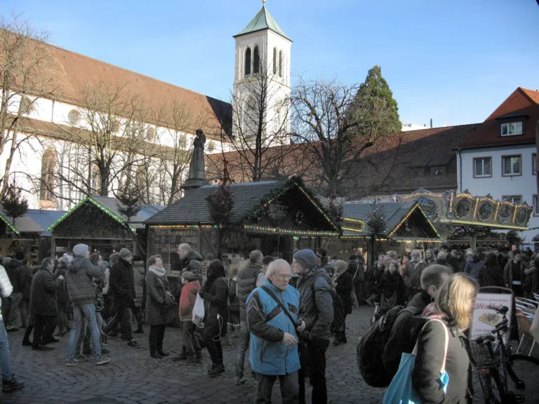 Christmas market stalls and visitors on Freiburg's Rathausplatz on a sunny winter day, with a church tower behind
