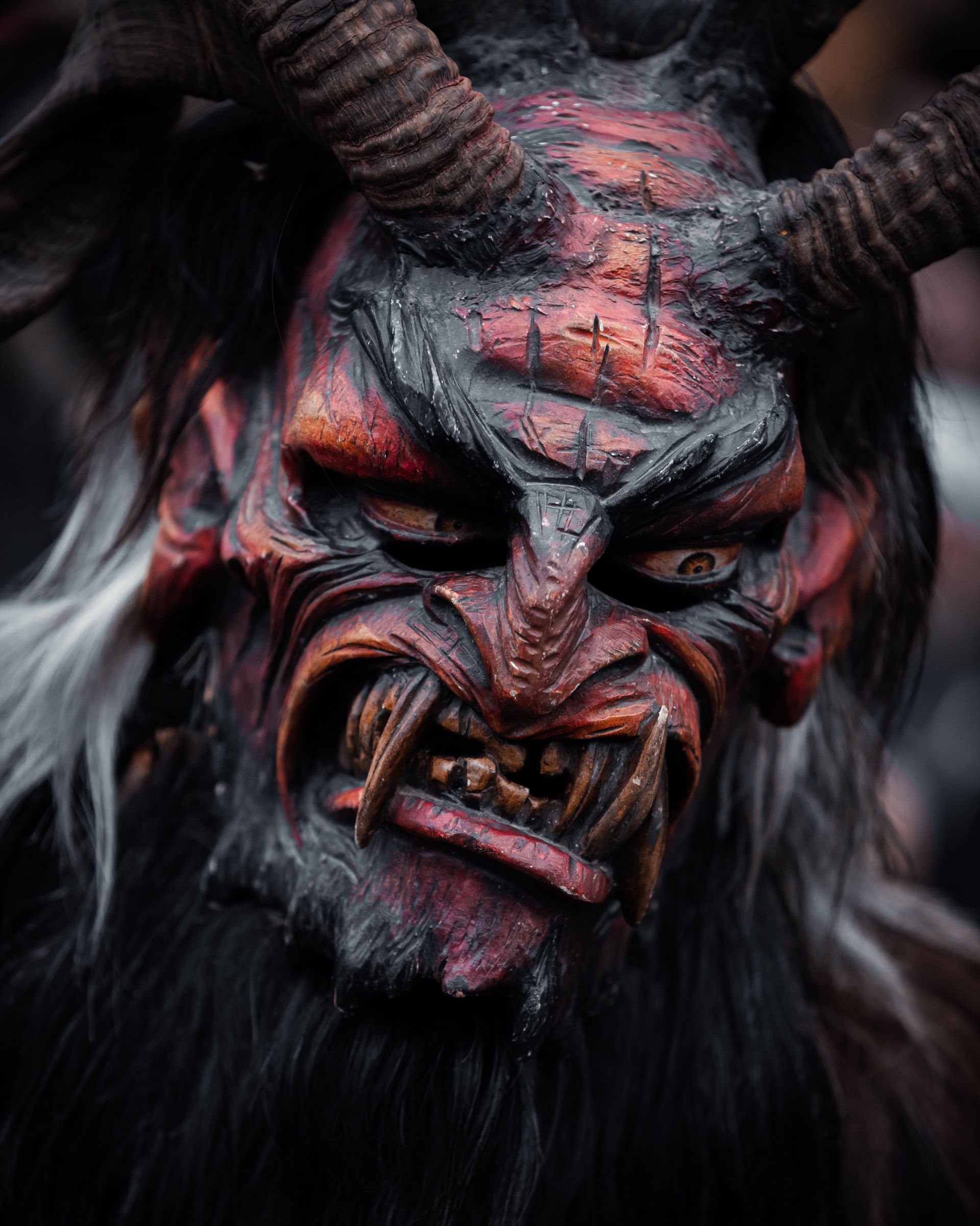 Close-up of a carved wooden Krampus mask painted red and black, with curved horns, fangs and a dark fur costume