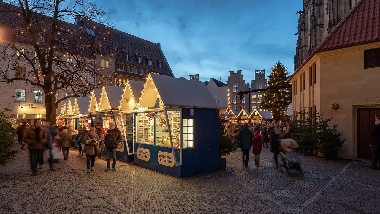 Beleuchtete Holzhütten eines Weihnachtsmarkts auf einem gepflasterten Altstadtplatz in der Abenddämmerung